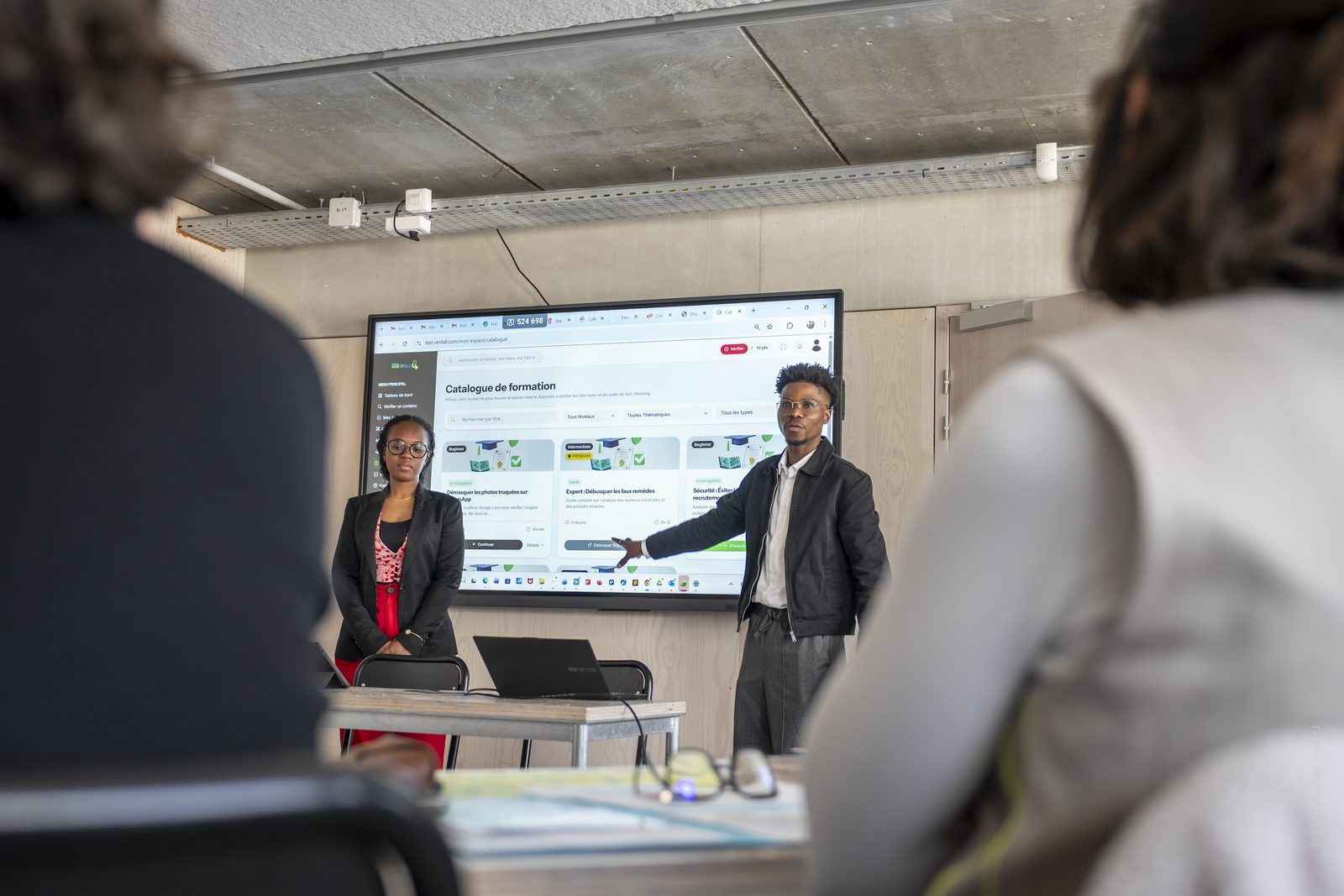 Démonstration de la plateforme Vérif'All Deux membres de l'équipe présentent le catalogue de formation de Vérif'All sur un grand écran, devant un public assis